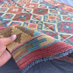 K&ouml;nnte beinhalten: Ein farbenfroher, gemusterter Teppich mit einer Mischung aus Rot-, Blau- und Beiget&ouml;nen. Der Teppich weist ein detailliertes Design mit Diamanten und quadratischen Formen auf. Der Rand des Teppichs ist sichtbar und zeigt einen Fransenrand.