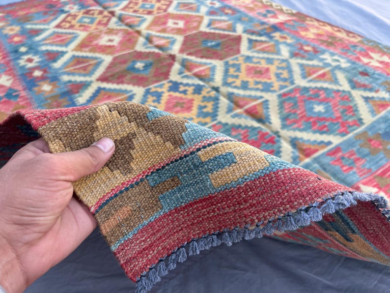 K&ouml;nnte beinhalten: Ein farbenfroher, gemusterter Teppich mit einer Mischung aus Rot-, Blau- und Beiget&ouml;nen. Der Teppich weist ein detailliertes Design mit Diamanten und quadratischen Formen auf. Der Rand des Teppichs ist sichtbar und zeigt einen Fransenrand.