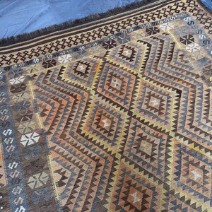 May include: A rectangular rug with a geometric diamond pattern in shades of brown, orange, and blue. The rug has a fringed edge and a border with repeating geometric shapes. The design is symmetrical and the rug appears to be made of woven material.