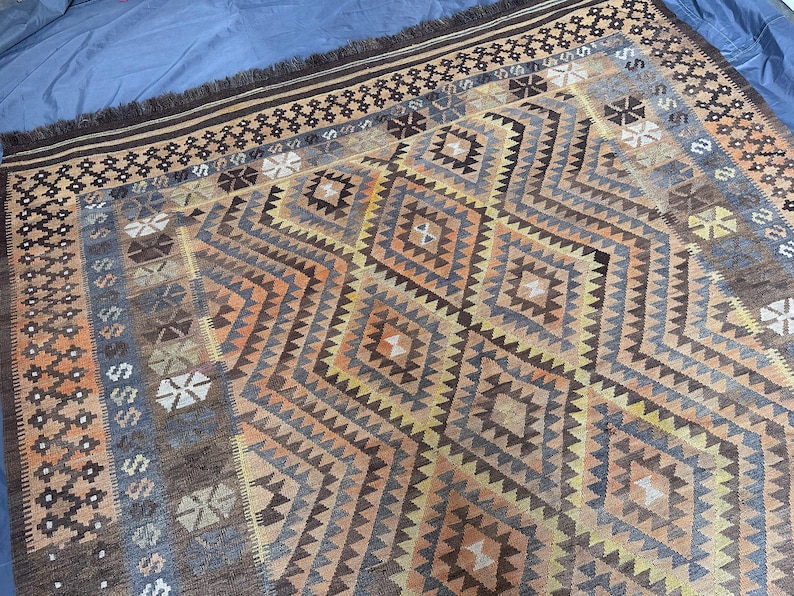 May include: A rectangular rug with a geometric diamond pattern in shades of brown, orange, and blue. The rug has a fringed edge and a border with repeating geometric shapes. The design is symmetrical and the rug appears to be made of woven material.