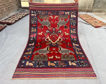 May include: A rectangular red rug with a detailed pattern featuring birds and geometric shapes. The rug has a blue border and a central design with symmetrical bird figures. The rug is likely hand-woven and adds a touch of cultural artistry.