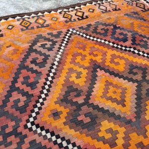 May include: A vibrant, geometric patterned rug in shades of orange, red, and navy blue. The rug features a repeating diamond and zigzag design, with a border of dark brown and black. This rug is a decorative floor covering.