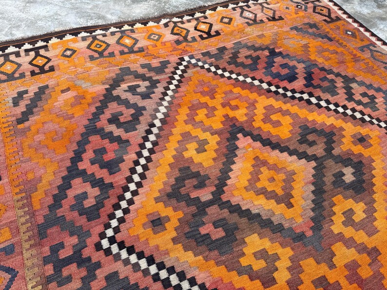 May include: A vibrant, geometric patterned rug in shades of orange, red, and navy blue. The rug features a repeating diamond and zigzag design, with a border of dark brown and black. This rug is a decorative floor covering.