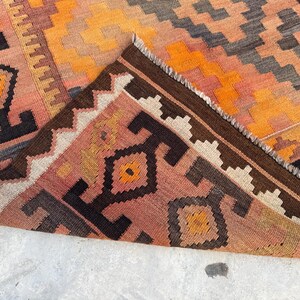 May include: A colorful, geometric patterned rug with a folded corner. The rug features a warm color palette of orange, peach, brown, and black. The design includes diamond and zig-zag patterns, with a fringed edge.