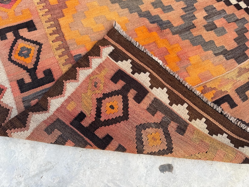 May include: A colorful, geometric patterned rug with a folded corner. The rug features a warm color palette of orange, peach, brown, and black. The design includes diamond and zig-zag patterns, with a fringed edge.