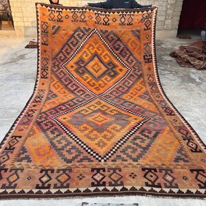 May include: Large, rectangular, hand-woven rug with a geometric design. The rug features a central diamond pattern in shades of orange, brown, and black, with intricate border details. The rug is displayed on a concrete surface.