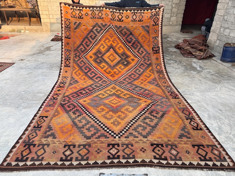 May include: Large, rectangular, hand-woven rug with a geometric design. The rug features a central diamond pattern in shades of orange, brown, and black, with intricate border details. The rug is displayed on a concrete surface.