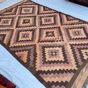 May include: A rectangular woven rug featuring a repeating geometric pattern of nested diamonds in shades of dark brown, light brown, and muted purple. The rug has a flat-weave texture and includes short, fringed edges on the short sides.
