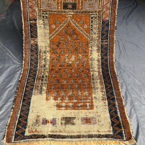 May include: A rectangular woven prayer rug featuring a central orange mihrab design with repeating dark geometric motifs. The border displays a blue and orange zigzag pattern, while the bottom section is cream-colored with fringe edges.