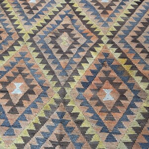 May include: A close-up of a woven rug with a geometric diamond pattern. The rug features a repeating design of diamonds and triangles in shades of brown, orange, blue, and yellow. The rug has a textured appearance.