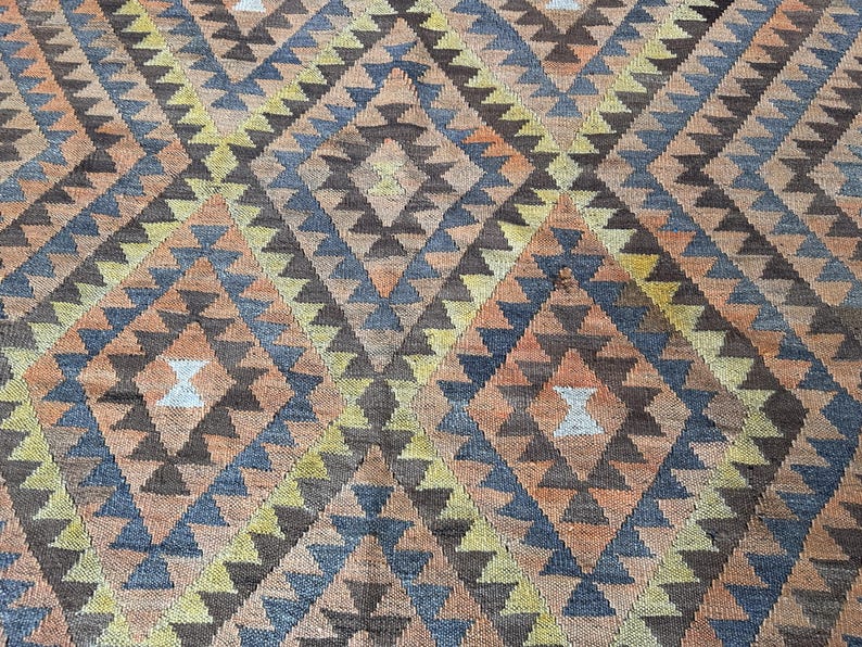 May include: A close-up of a woven rug with a geometric diamond pattern. The rug features a repeating design of diamonds and triangles in shades of brown, orange, blue, and yellow. The rug has a textured appearance.