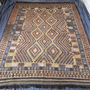 May include: A rectangular, hand-woven rug with a geometric diamond pattern in shades of brown, tan, and yellow. The rug has a dark brown border with fringe along the edges. The central design features repeating diamond and triangle shapes.