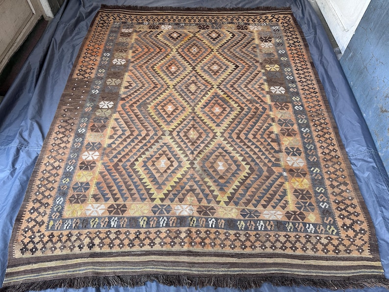 May include: A rectangular, hand-woven rug with a geometric diamond pattern in shades of brown, tan, and yellow. The rug has a dark brown border with fringe along the edges. The central design features repeating diamond and triangle shapes.