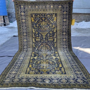 May include: A large, rectangular rug with an intricate, geometric design. The rug features a dark blue background with golden yellow and cream-colored patterns. The border has a repeating floral motif. The rug is displayed outdoors.