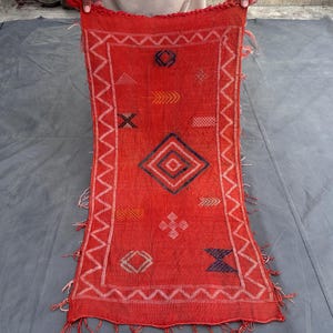 Puede incluir: Textil rectangular rojo con bordes en zigzag blancos y patrones geométricos. El diseño incluye motivos de diamantes, flechas y cruces en azul, naranja y blanco. Flecos adornan los extremos del textil.
