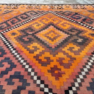 May include: A large, rectangular rug with a geometric pattern in shades of orange, red, brown, and navy blue. The central design features a diamond shape with smaller diamond and square patterns. The rug has a woven texture and a border with a repeating pattern.