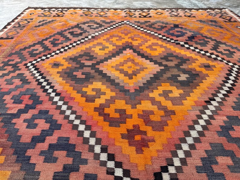 May include: A large, rectangular rug with a geometric pattern in shades of orange, red, brown, and navy blue. The central design features a diamond shape with smaller diamond and square patterns. The rug has a woven texture and a border with a repeating pattern.