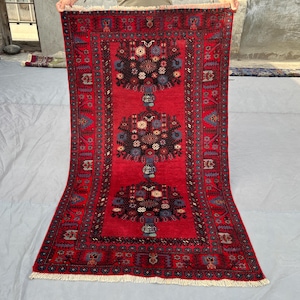 May include: A rectangular red rug with a central design of three stylized floral arrangements in black, blue, and white. The rug has a detailed border with repeating patterns and a fringed edge. The rug is displayed against a neutral background.