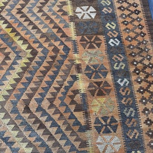 May include: A close-up of a woven rug with a geometric pattern. The rug features a diamond and triangle design in shades of brown, orange, yellow, and blue. The border has a repeating pattern of shapes and symbols.