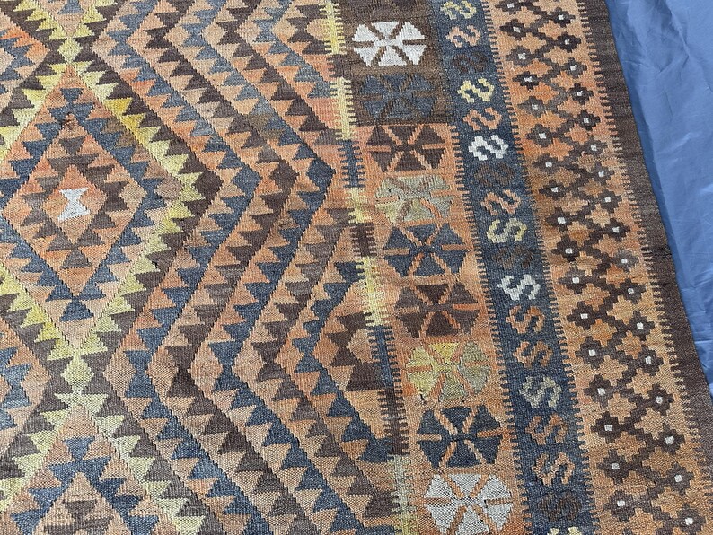 May include: A close-up of a woven rug with a geometric pattern. The rug features a diamond and triangle design in shades of brown, orange, yellow, and blue. The border has a repeating pattern of shapes and symbols.