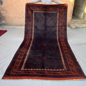 Puede incluir: Una alfombra rectangular de color burdeos oscuro con un intrincado patrón geométrico. La alfombra presenta un campo central con un diseño repetitivo, enmarcado por bordes en tonos naranja, marrón y crema. La alfombra se exhibe sobre una superficie gris claro.