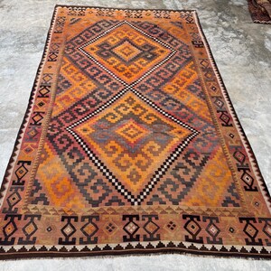 May include: A rectangular, hand-woven rug with a geometric pattern in shades of orange, brown, and gray. The central design features a diamond shape with smaller diamond and zigzag patterns. The rug has a dark brown border.
