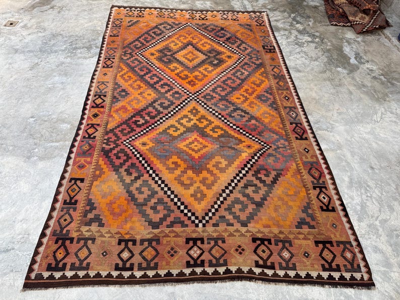 May include: A rectangular, hand-woven rug with a geometric pattern in shades of orange, brown, and gray. The central design features a diamond shape with smaller diamond and zigzag patterns. The rug has a dark brown border.