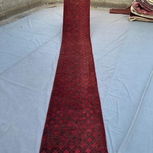 Könnte beinhalten: Ein langer, rechteckiger roter Teppich mit einem sich wiederholenden geometrischen Muster in Dunkelgrün und Schwarz. Der Teppich wird hochgehalten, um seine Länge und sein kompliziertes Design zu zeigen. Der Teppich scheint aus gewebtem Material zu bestehen.