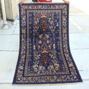 Puede incluir: Una alfombra rectangular con un fondo azul profundo y patrones florales y botánicos intrincados en tonos crema, marrón y rojo oscuro. La alfombra tiene un borde decorativo ancho y flecos cortos en los bordes superior e inferior.