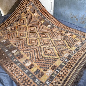 May include: A rectangular, hand-woven rug with a geometric pattern in shades of brown, tan, and gold. The rug features diamond and zigzag designs, with a fringed edge. The overall aesthetic is rustic and traditional.