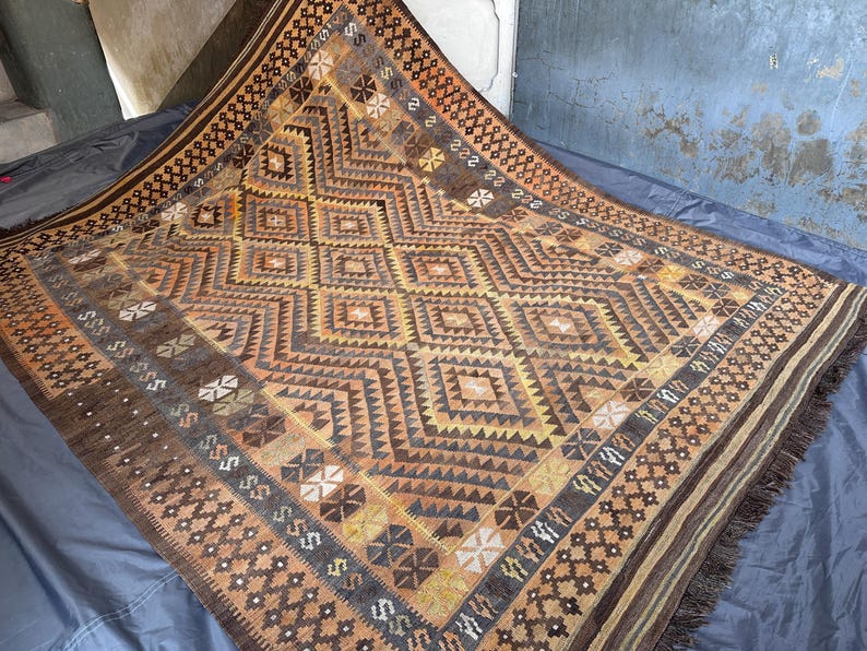 May include: A rectangular, hand-woven rug with a geometric pattern in shades of brown, tan, and gold. The rug features diamond and zigzag designs, with a fringed edge. The overall aesthetic is rustic and traditional.
