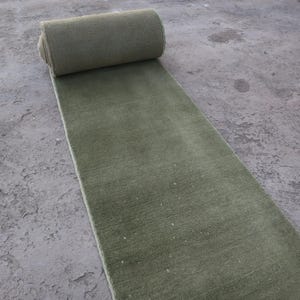 Può includere: Un tappeto verde oliva arrotolato su una superficie di cemento. Il tappeto è rettangolare e ha un aspetto morbido e strutturato. La parte arrotolata è in alto, con la sezione srotolata che si estende verso il basso. Il colore è uniforme.