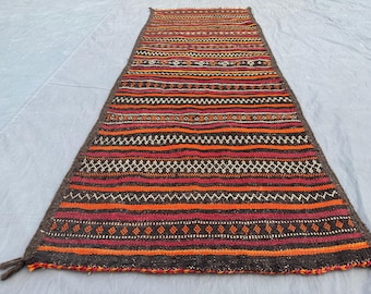 May include: A rectangular, hand-woven rug with a geometric pattern. The rug features horizontal stripes in shades of orange, red, and brown, with white and black accents. The rug has a dark brown border and tassels on one end.
