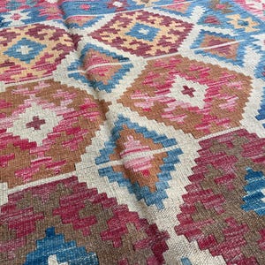 K&ouml;nnte beinhalten: Nahaufnahme eines gewebten Teppichs mit geometrischem Muster. Der Teppich zeigt Rauten in Blau-, Rosa-, Braun- und Beiget&ouml;nen. Der Teppich hat ein strukturiertes Aussehen, was auf eine handgefertigte Qualit&auml;t hindeutet.