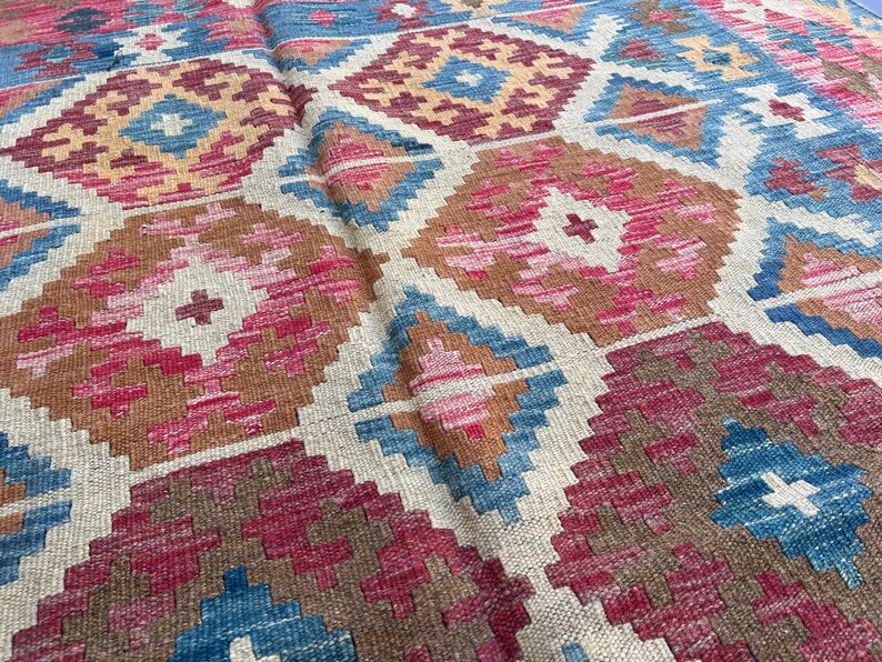 K&ouml;nnte beinhalten: Nahaufnahme eines gewebten Teppichs mit geometrischem Muster. Der Teppich zeigt Rauten in Blau-, Rosa-, Braun- und Beiget&ouml;nen. Der Teppich hat ein strukturiertes Aussehen, was auf eine handgefertigte Qualit&auml;t hindeutet.