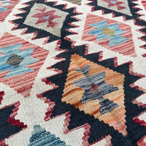 Op de afbeelding: Een close-up van een kleurrijk tapijt met een geometrisch patroon. Het tapijt heeft een verscheidenheid aan kleuren, waaronder rood, blauw, oranje en zwart. Het patroon bestaat uit herhalende ruiten en driehoeken.
