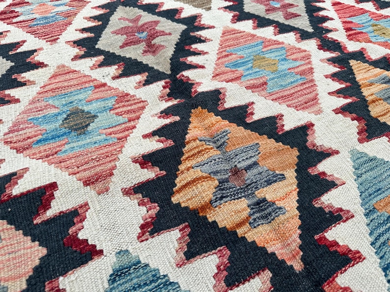 Op de afbeelding: Een close-up van een kleurrijk tapijt met een geometrisch patroon. Het tapijt heeft een verscheidenheid aan kleuren, waaronder rood, blauw, oranje en zwart. Het patroon bestaat uit herhalende ruiten en driehoeken.