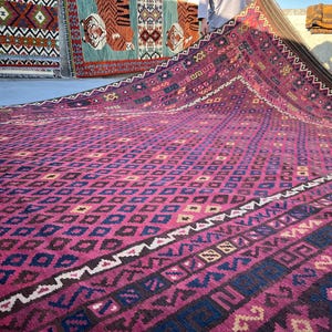 May include: A large, rectangular rug with a repeating diamond pattern in shades of pink and blue. The rug has a border with geometric designs and a dark brown edge. Other rugs are visible in the background.