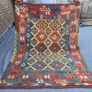 K&ouml;nnte beinhalten: Ein rechteckiger, handgewebter Teppich mit geometrischem Muster. Der Teppich weist ein zentrales Design aus Rauten und anderen Formen in Rot-, Blau-, Beige- und Braunt&ouml;nen auf. Der Teppich wird von einer Person gehalten, was seine Gr&ouml;&szlig;e und komplizierten Details zeigt.