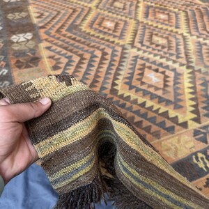 May include: A hand-woven rug with a geometric diamond pattern in shades of brown, orange, and beige. The rug's edge is folded over, revealing a striped border with fringe. The rug's design features repeating diamond and triangular shapes.