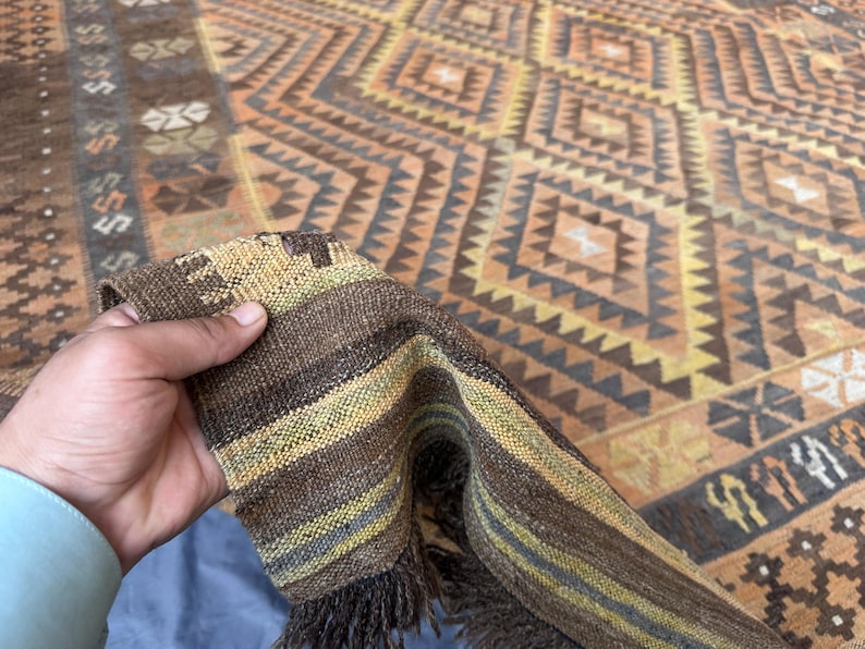 May include: A hand-woven rug with a geometric diamond pattern in shades of brown, orange, and beige. The rug's edge is folded over, revealing a striped border with fringe. The rug's design features repeating diamond and triangular shapes.
