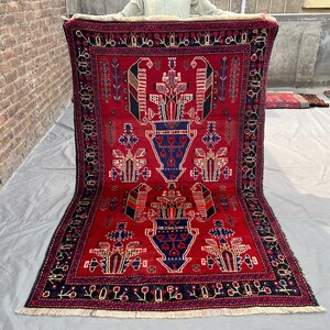 Könnte beinhalten: Ein rechteckiger roter Teppich mit einem detaillierten geometrischen Muster. Der Teppich hat ein zentrales Design aus stilisierten Vasen und floralen Motiven in Blau, Schwarz und Creme. Der Rand ist dunkel mit komplizierten Mustern.