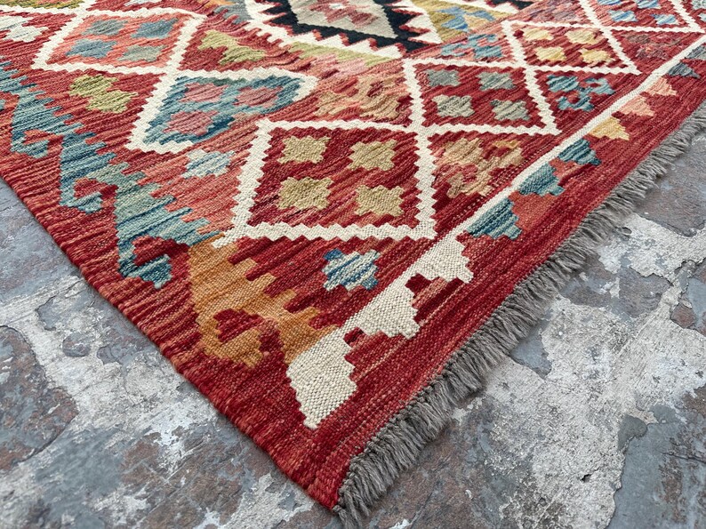 Op de afbeelding: Een rood, oranje en blauw patroonteppich met een franje rand. Het tapijt heeft een geometrisch ontwerp met ruitvormen en ingewikkelde details.