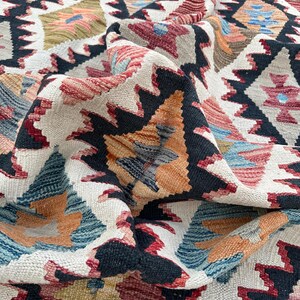 Op de afbeelding: Een close-up van een kleurrijk geweven tapijt met een geometrisch patroon. Het tapijt is gemaakt van wol en heeft een zacht, getextureerd gevoel. De kleuren zijn levendig en omvatten tinten rood, blauw, groen en geel.