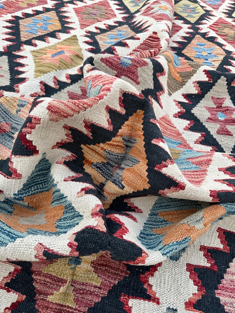 Op de afbeelding: Een close-up van een kleurrijk geweven tapijt met een geometrisch patroon. Het tapijt is gemaakt van wol en heeft een zacht, getextureerd gevoel. De kleuren zijn levendig en omvatten tinten rood, blauw, groen en geel.