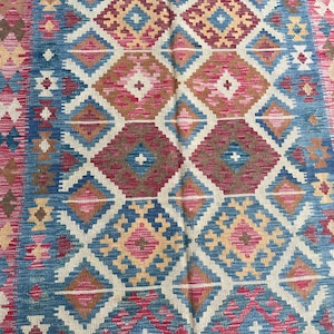 K&ouml;nnte beinhalten: Ein farbenfroher Teppich mit geometrischem Muster. Der Teppich zeigt ein sich wiederholendes Rautenmuster in Blau-, Rot-, Braun- und Cremet&ouml;nen. Der Rand des Teppichs hat ein Zickzackmuster. Der Teppich besteht aus einem gewebten Material.