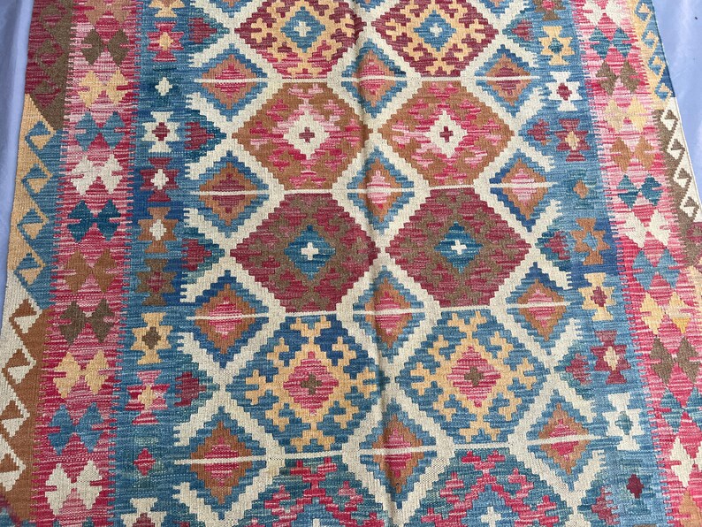 K&ouml;nnte beinhalten: Ein farbenfroher Teppich mit geometrischem Muster. Der Teppich zeigt ein sich wiederholendes Rautenmuster in Blau-, Rot-, Braun- und Cremet&ouml;nen. Der Rand des Teppichs hat ein Zickzackmuster. Der Teppich besteht aus einem gewebten Material.