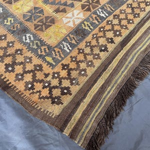 May include: A corner of a hand-woven rug with a geometric pattern in shades of brown, tan, yellow, and gray. The rug has a fringed edge and a detailed design, showcasing traditional craftsmanship. The rug's texture and color palette create a warm, inviting aesthetic.