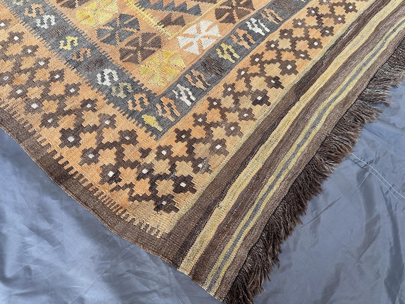 May include: A corner of a hand-woven rug with a geometric pattern in shades of brown, tan, yellow, and gray. The rug has a fringed edge and a detailed design, showcasing traditional craftsmanship. The rug's texture and color palette create a warm, inviting aesthetic.