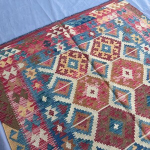 K&ouml;nnte beinhalten: Ein farbenfroher Teppich mit geometrischem Muster in Rot-, Blau-, Beige- und Goldt&ouml;nen. Das Design des Teppichs zeigt sich in sich wiederholenden Diamanten und sechseckigen Formen, die ein strukturiertes Aussehen erzeugen. Der Teppich ist wahrscheinlich aus gewebtem Material.